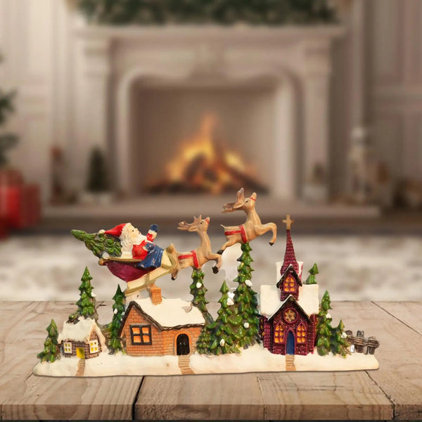 Resin Christmas village figurine with Santa and reindeer flying over snowy houses and trees on a wooden surface.